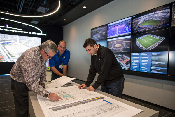 Professional team reviewing architectural plans with stadium displays showing sports venues in background